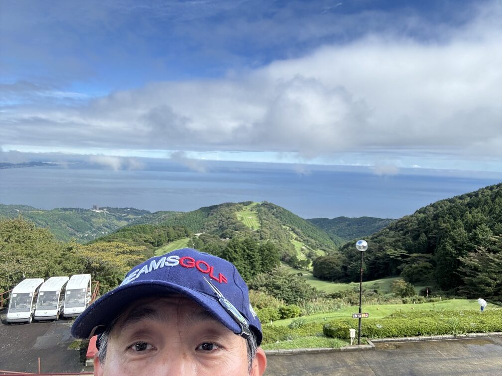 ゴルフ場の山間から広がる美しい海と青空の風景を背景に、キャップをかぶった男性が写る